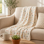 A cream-colored muslin throw blanket with a small multicolor heart pattern is draped over a beige armchair in a cozy living room setting.