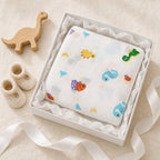 A Kiddospot white muslin baby blanket with colorful dinosaur prints, presented in a white gift box with white tissue paper. The gift box is styled on a beige fabric background with a wooden dinosaur toy and cream-colored knit booties.