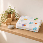 A folded Kiddospot white muslin baby blanket with colorful cartoon sea creatures and fish prints sits on a light wood shelf next to a wooden dinosaur toy, a potted plant, and a smooth stone.