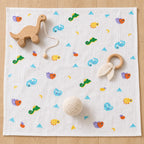 A square Kiddospot white muslin baby blanket with a colorful cartoon print of fish, dinosaurs, and suns, styled with wooden and fabric baby toys on a light wood floor.