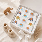 A folded Kiddospot white muslin baby blanket with a colorful elephant print sits in an open white gift box tied with a white ribbon. The blanket features elephants in blue, purple, green, and gray with orange centers.
