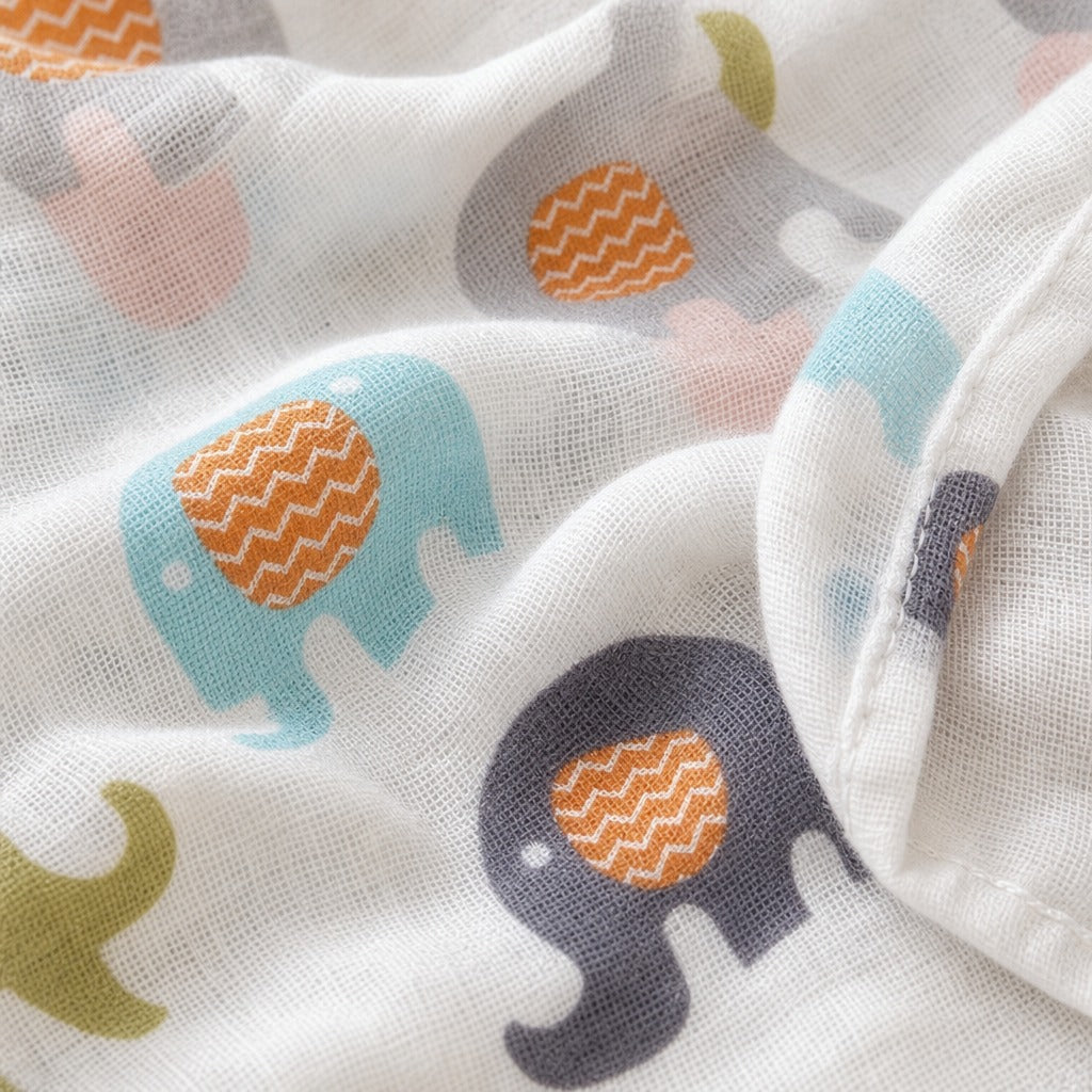 Close-up of a white Kiddospot muslin blanket with a playful elephant print featuring light blue, dark navy, pink, gray, and olive green elephants with orange chevron-patterned trunks.
