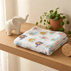A folded Kiddospot white muslin baby swaddle blanket with a colorful elephant print is displayed on a light wood shelf next to a white ceramic elephant figurine, a potted green plant, and a wooden star-shaped coaster.