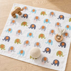 A Kiddospot white muslin baby blanket with a repeating pattern of colorful elephants, styled on a light wood floor with baby toys including a wooden elephant toy, a wooden teething ring, and a cream knit ball.