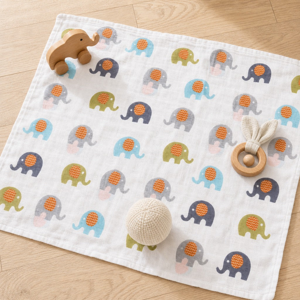 A Kiddospot white muslin baby blanket with a repeating pattern of colorful elephants, styled on a light wood floor with baby toys including a wooden elephant toy, a wooden teething ring, and a cream knit ball.