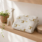 A folded Kiddospot white muslin blanket with a yellow and brown giraffe pattern sits on a light wood shelf next to a white giraffe figurine, a potted plant, and a wood slice.