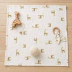 A square Kiddospot white muslin baby blanket with a repeating gold giraffe pattern, styled on a light wood floor with a wooden giraffe teether, a wooden ring rattle with a white fabric bow, and a cream-colored knit ball.