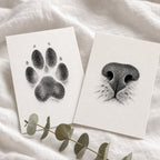 Two white art prints laid on a soft white fabric background, one showing a black paw print and the other a black cat nose illustration, with a eucalyptus sprig in front.