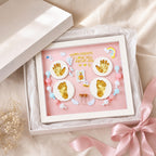 Kiddospot baby keepsake framed shadow box gift set displayed in an open white box with a pink insert. The frame contains gold baby foot and handprints, white circular bases, pastel beads, small angel wings, and the text 'HAPPY 100 DAYS I LOVE YOU ABCMS-2165 W 19 Q' on a soft pink background.