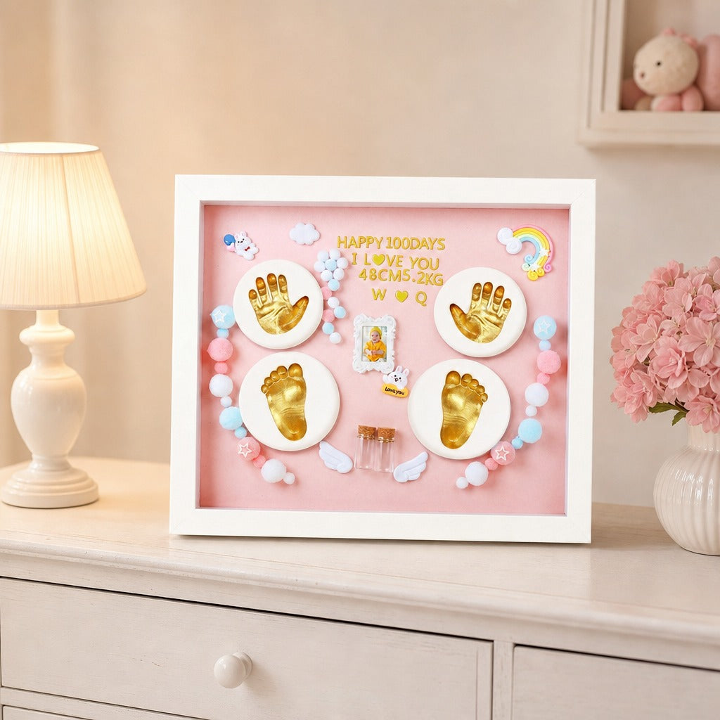 Kiddospot framed baby keepsake shadow box displayed on a light-colored dresser next to a lamp and pink flowers. The white frame contains a pink background with gold baby footprints and handprints, small pastel beads, and a central photo slot.