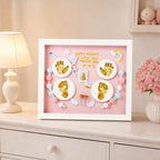 Kiddospot framed baby keepsake shadow box displayed on a light-colored dresser next to a lamp and pink flowers. The white frame contains a pink background with gold baby footprints and handprints, small pastel beads, and a central photo slot.