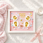 Kiddospot framed baby keepsake shadow box displayed on a textured white fabric surface with a pink blanket, candle, and ribbon tag nearby. The frame contains a pink background with gold foil baby footprints and handprints, small pastel beads, angel wings, a tiny photo slot, and the text 'HAPPY 100 DAYS I LOVE YOU 48CM5.