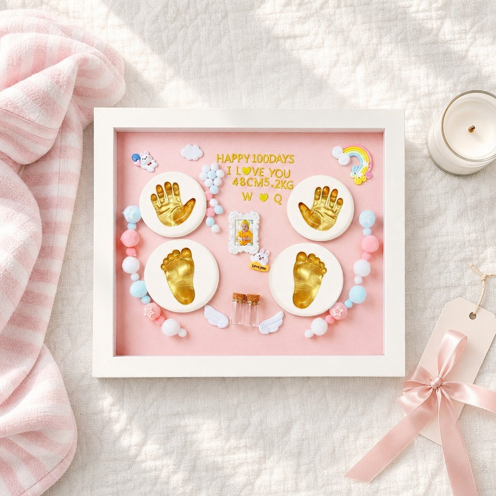 Kiddospot framed baby keepsake shadow box displayed on a textured white fabric surface with a pink blanket, candle, and ribbon tag nearby. The frame contains a pink background with gold foil baby footprints and handprints, small pastel beads, angel wings, a tiny photo slot, and the text 'HAPPY 100 DAYS I LOVE YOU 48CM5.