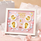 Kiddospot framed baby keepsake display with a white frame and pink backing, featuring gold footprints and handprints, pastel beads, small wings, a tiny photo frame, and gold lettering that reads 'HAPPY 100DAYS I LOVE YOU 48CM.5.