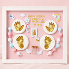 Kiddospot framed 100-day baby milestone shadow box display on a pink background. The white frame contains gold baby handprints and footprints on round white plaques, a small photo frame, pastel beads, white angel wings, and gold text reading 'HAPPY 100 DAYS I LOVE YOU 48CM5.