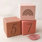 Kiddospot's wooden milestone blocks stacked in a pyramid arrangement on a light beige surface. The bottom-left block is terracotta pink with the number 5, the bottom-right block is coral pink with the number 7, and the top block is pale pink with a dark brown rainbow design.