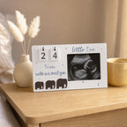 A white baby milestone plaque styled on a wooden bedside table, featuring a flip calendar showing 24 weeks, blue script text, elephant silhouettes, and a black-and-white ultrasound photo. Decorative pampas grass in a beige vase and a yellow cup are in the background.