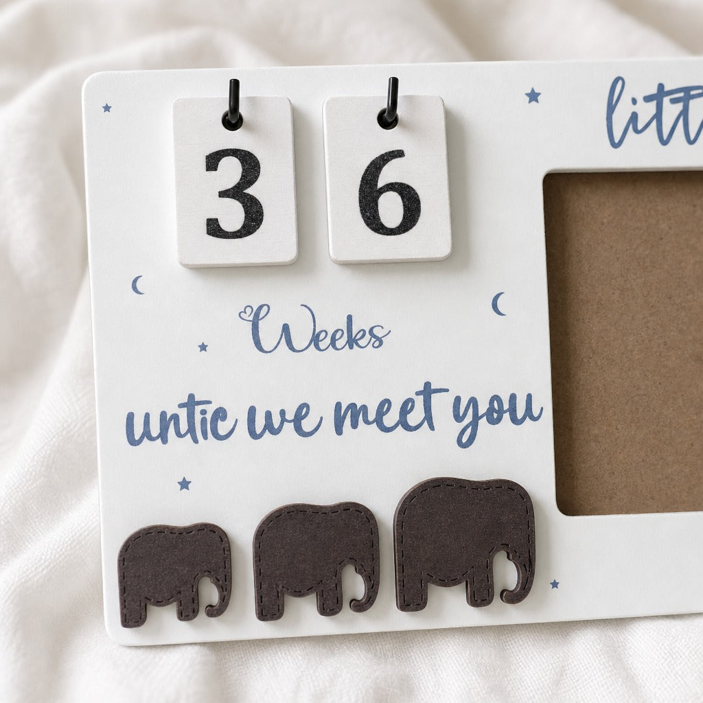 A white baby milestone calendar plaque with wooden flip days showing 3 and 6, blue script text reading 'Weeks untic we meet you,' small blue moon and star accents, and three dark brown elephant cutouts along the bottom. It is photographed on a soft white fabric background.
