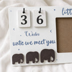 A white baby milestone calendar plaque with wooden flip days showing 3 and 6, blue script text reading 'Weeks untic we meet you,' small blue moon and star accents, and three dark brown elephant cutouts along the bottom. It is photographed on a soft white fabric background.