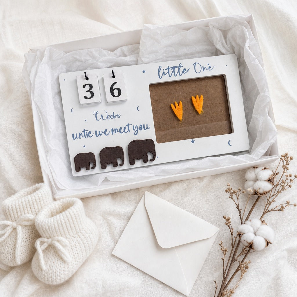 A white personalized baby photo frame gift box on a soft white fabric background, styled with cream knit baby booties, a white envelope, and dried cotton stems. The frame has a brown insert with a pair of orange bootie prints and the text 'little One' and '36 Weeks until we meet you'.