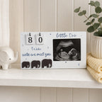 A white baby keepsake plaque on a white shelf, featuring a flip-day calendar showing 80 days, the text 'little One' and 'Weds untie we meet you', three elephant silhouettes, and a black-and-white ultrasound photo. Decorative props include a small white elephant figurine, a potted plant, and a folded cream blanket.