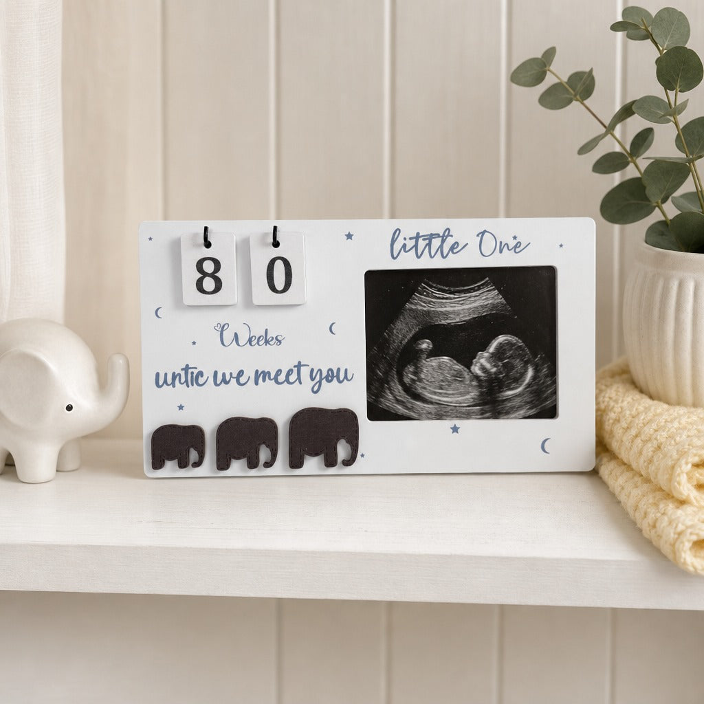 A white baby keepsake plaque on a white shelf, featuring a flip-day calendar showing 80 days, the text 'little One' and 'Weds untie we meet you', three elephant silhouettes, and a black-and-white ultrasound photo. Decorative props include a small white elephant figurine, a potted plant, and a folded cream blanket.