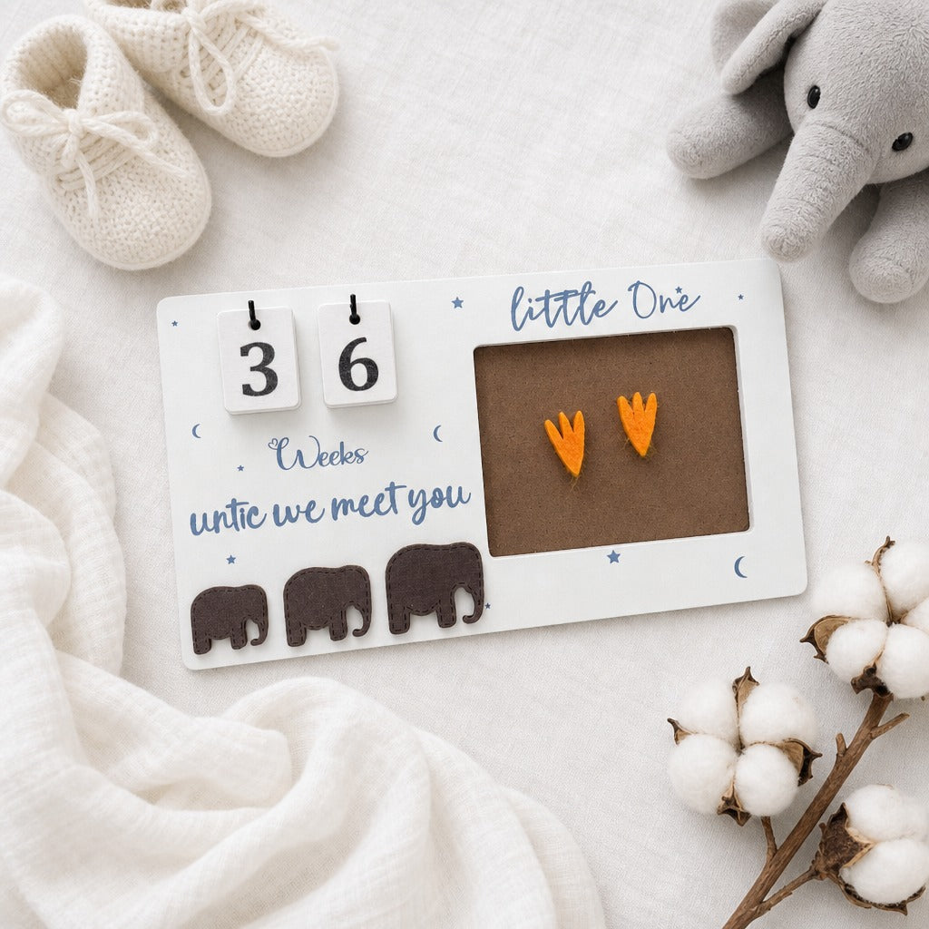 A baby milestone calendar plaque on a white fabric background, styled with cream knit baby shoes, a gray plush elephant, and cotton bolls. The white rectangular plaque has a flip-day calendar showing 36 weeks until they meet you, blue script text, small moon and star details, three dark brown elephant silhouettes, and a brown photo window holding two orange bootie-shaped stickers.