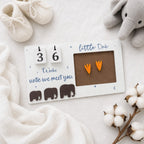 A baby milestone calendar plaque on a white fabric background, styled with cream knit baby shoes, a gray plush elephant, and cotton bolls. The white rectangular plaque has a flip-day calendar showing 36 weeks until they meet you, blue script text, small moon and star details, three dark brown elephant silhouettes, and a brown photo window holding two orange bootie-shaped stickers.