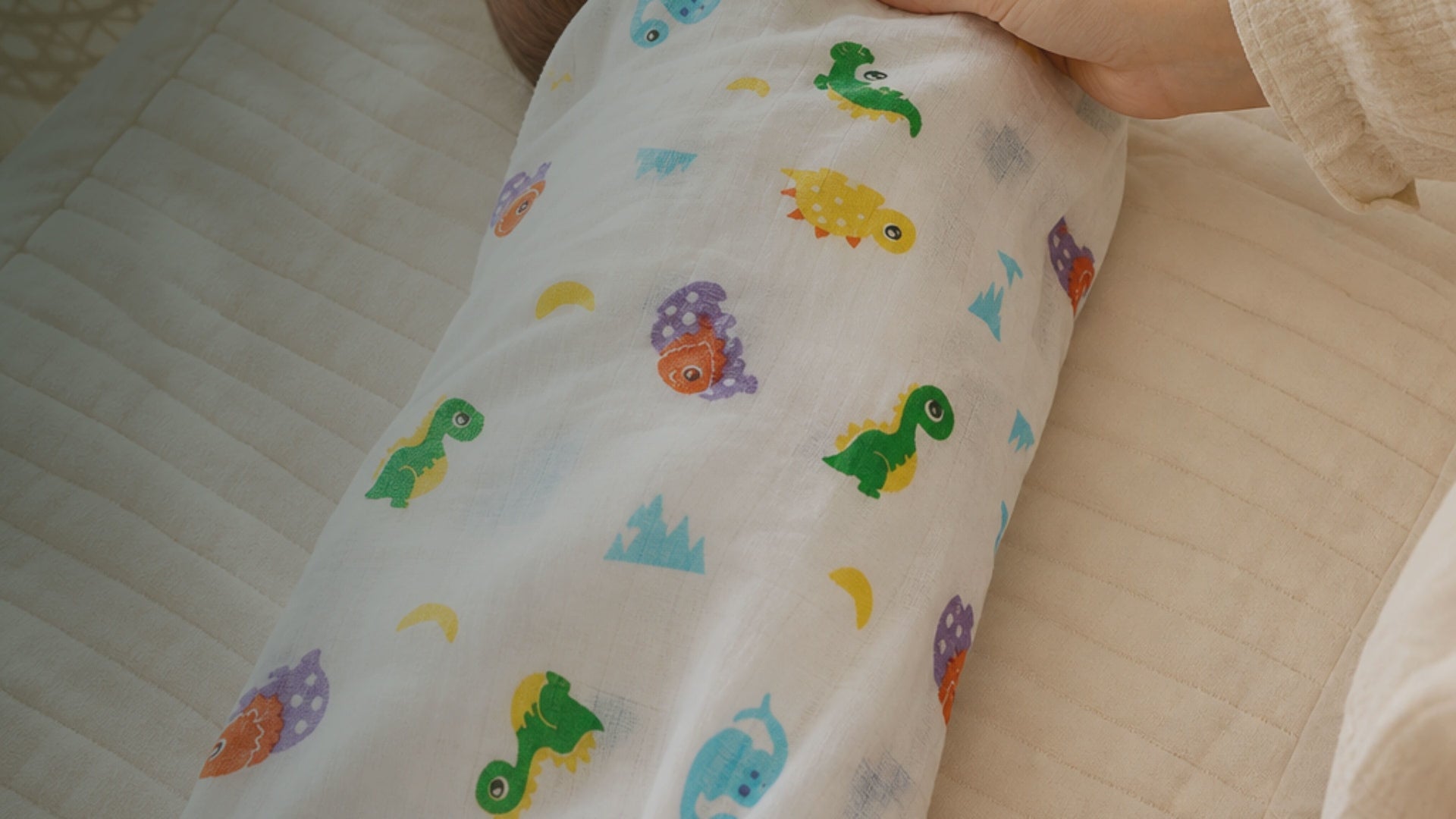 Kiddospot GOTS Certified Organic Cotton Muslin Swaddle in Dinosaur Print — crisp white double-gauze muslin with colourful cartoon dinosaurs in kelly green, sky blue, marigold yellow, orange and lavender purple, with small yellow crescent moons and pale blue mountain accents. 120x120cm. Breathable open weave for safe sleep temperature regulation. Bold, playful, gender-neutral design. Multi-use: swaddle, pram shade, nursing cover, tummy time mat. Free AU shipping. kiddospot.com