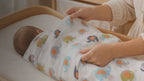 Kiddospot GOTS Certified Organic Cotton Muslin Swaddle in Elephant Print — white double-gauze muslin with colourful elephant motifs in sky blue, charcoal grey, sage green and dusty pink, featuring distinctive orange chevron ears. 120x120cm. Breathable open weave for safe temperature regulation. Multi-use: swaddle, pram shade, nursing cover, tummy time mat. Gets softer with every wash. Free AU shipping. kiddospot.com