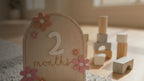 Kiddospot Wooden Baby Milestone Cards — set of 12 reusable arch-shaped natural birch discs with raised white 3D numbers, laser-engraved cursive "months" text, and 3D wooden daisy flowers in dusty pink and mustard yellow. Gender-neutral boho nursery aesthetic. Perfect monthly baby photo prop and baby shower gift. Free AU shipping. kiddospot.com