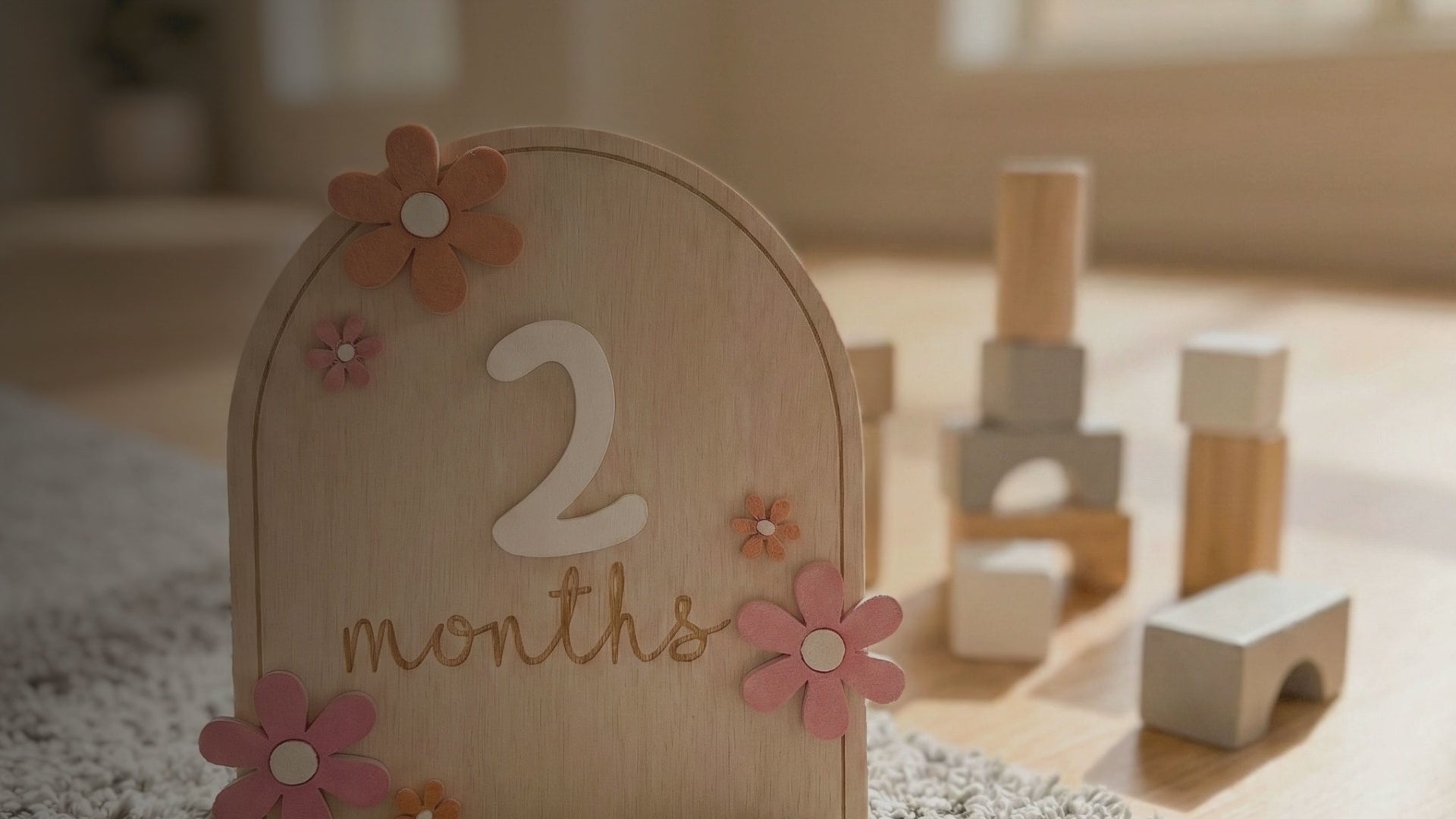 Kiddospot Wooden Baby Milestone Cards — set of 12 reusable arch-shaped natural birch discs with raised white 3D numbers, laser-engraved cursive "months" text, and 3D wooden daisy flowers in dusty pink and mustard yellow. Gender-neutral boho nursery aesthetic. Perfect monthly baby photo prop and baby shower gift. Free AU shipping. kiddospot.com