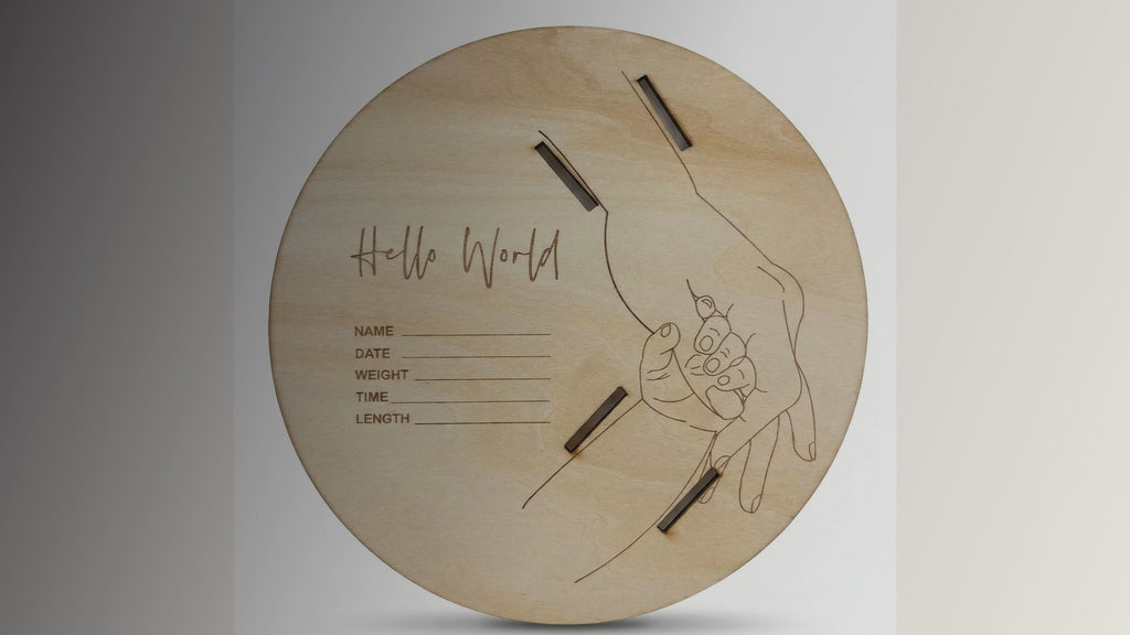 Kiddospot Wooden Baby Announcement Sign and Hospital Bracelet Holder — circular natural birch disc with laser-engraved "Hello World" cursive text, fine-line adult and newborn hands illustration, engraved fields for name, date, time, weight and length, and four laser-cut rectangular slots to hold both mum's and baby's hospital wristbands. AS/NZS certified. Newborn photo prop and permanent birth keepsake. Free AU shipping. kiddospot.com