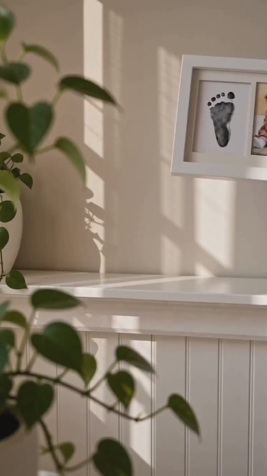 Vertical video of parent hands placing a white triple-window inkless newborn handprint and footprint keepsake frame on a nursery shelf