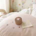 Kiddospot small round wooden tooth fairy box sits on a soft pink blanket on a bed, next to a handwritten note that says 'Dear Tooth Fairy.' The box lid is decorated with an illustration of a tooth fairy and the words 'Tooth Fairy Box.