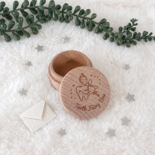 Kiddospot round wooden tooth fairy box with its lid open, engraved with a tooth fairy illustration and the words 'Tooth Fairy Box.' It is placed on a white faux-fur surface with scattered silver star confetti, a small cream envelope, and green decorative foliage in the background.
