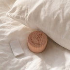 Kiddospot small round wooden keepsake box with a fairy engraving sits on white bedding next to a folded white card, with a white pillow in the background.