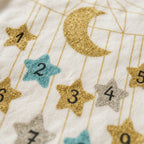 Close-up of a white fabric surface printed with a gold crescent moon and numbered glitter stars in gold, silver, and light blue.