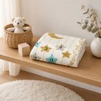 A folded baby blanket with a celestial pattern of gold stars, silver stars, a gold crescent moon, and colorful numbered star shapes sits on a wooden shelf next to a woven basket with a teddy bear and a small wooden block.