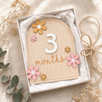 Kiddospot wooden 3-month milestone plaque with a white number 3 and the word 'months' in script, decorated with pink and yellow wooden flowers, presented in a white gift box on a beige fabric background with dried flowers and eucalyptus leaves.
